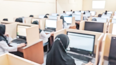 A computer lab filled with several individuals seated at workstations, focusing on computer screens. Each person is seated in a wooden cubicle, providing a semi-private work environment. The setting suggests a formal environment, like a school or office.
