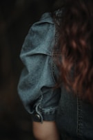 A close-up of a person wearing a denim jacket. The fabric appears textured, with visible seams and a buttoned cuff. The focus is on the jacket's shoulder and sleeve area. Brown, wavy hair is partially visible flowing alongside the jacket.