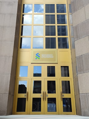 A tall building entrance with large yellow-framed glass doors and windows. The logo of Standard Chartered is prominently displayed above the doors. The reflection in the windows shows a blue sky with white clouds.