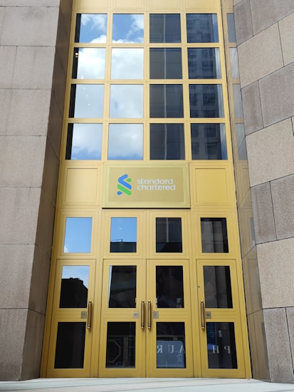 A tall building entrance with large yellow-framed glass doors and windows. The logo of Standard Chartered is prominently displayed above the doors. The reflection in the windows shows a blue sky with white clouds.