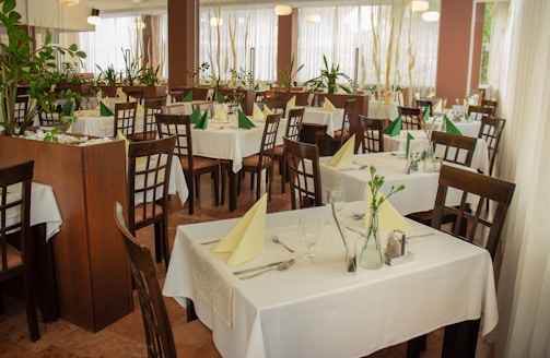 The image depicts a well-arranged dining area in a restaurant with wooden chairs and tables covered with white tablecloths. Each table is set with cutlery, glasses, and folded napkins, either in green or yellow. There are several potted plants around, adding a natural and refreshing touch to the decor. Large windows with sheer white curtains allow soft natural light to fill the space.