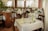 The image depicts a well-arranged dining area in a restaurant with wooden chairs and tables covered with white tablecloths. Each table is set with cutlery, glasses, and folded napkins, either in green or yellow. There are several potted plants around, adding a natural and refreshing touch to the decor. Large windows with sheer white curtains allow soft natural light to fill the space.