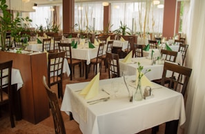 The image depicts a well-arranged dining area in a restaurant with wooden chairs and tables covered with white tablecloths. Each table is set with cutlery, glasses, and folded napkins, either in green or yellow. There are several potted plants around, adding a natural and refreshing touch to the decor. Large windows with sheer white curtains allow soft natural light to fill the space.