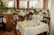 The image depicts a well-arranged dining area in a restaurant with wooden chairs and tables covered with white tablecloths. Each table is set with cutlery, glasses, and folded napkins, either in green or yellow. There are several potted plants around, adding a natural and refreshing touch to the decor. Large windows with sheer white curtains allow soft natural light to fill the space.
