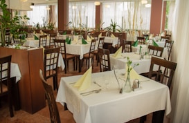 The image depicts a well-arranged dining area in a restaurant with wooden chairs and tables covered with white tablecloths. Each table is set with cutlery, glasses, and folded napkins, either in green or yellow. There are several potted plants around, adding a natural and refreshing touch to the decor. Large windows with sheer white curtains allow soft natural light to fill the space.