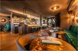 A modern and elegantly designed bar interior with warm lighting that creates a cozy atmosphere. The bar is stocked with various bottles and glasses hanging above. Plush green and brown seating, coupled with wooden tables, enhances the luxurious vibe.