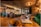 A modern and elegantly designed bar interior with warm lighting that creates a cozy atmosphere. The bar is stocked with various bottles and glasses hanging above. Plush green and brown seating, coupled with wooden tables, enhances the luxurious vibe.