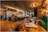 A modern and elegantly designed bar interior with warm lighting that creates a cozy atmosphere. The bar is stocked with various bottles and glasses hanging above. Plush green and brown seating, coupled with wooden tables, enhances the luxurious vibe.
