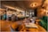 A modern and elegantly designed bar interior with warm lighting that creates a cozy atmosphere. The bar is stocked with various bottles and glasses hanging above. Plush green and brown seating, coupled with wooden tables, enhances the luxurious vibe.