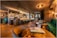 A modern and elegantly designed bar interior with warm lighting that creates a cozy atmosphere. The bar is stocked with various bottles and glasses hanging above. Plush green and brown seating, coupled with wooden tables, enhances the luxurious vibe.