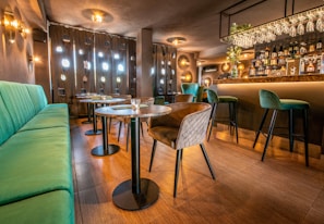 A well-designed bar interior featuring a mix of modern and cozy elements. The space includes green cushioned benches and chairs arranged around wooden tables. The lighting is warm and ambient, with decorative circular fixtures on the walls. The bar area is well-stocked, with glassware hanging above and various bottles on display, complemented by a floral arrangement.