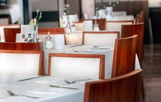 A stylish restaurant interior featuring elegant, wooden chairs with white cushions surrounding tables covered with crisp white tablecloths. Each table is set with silverware and decorated with small vases containing delicate flowers. The setting conveys a neat and upscale dining atmosphere.