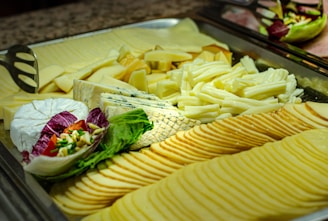 A platter showcasing various types of cheese arranged neatly, including sliced and block varieties. There is a decorative addition of leafy greens and purple lettuce with a garnish of chives and chopped vegetables. The setup is on a metal tray with serving utensils.