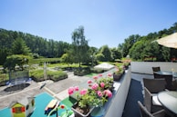 A spacious outdoor terrace featuring wicker furniture and a row of vibrant pink flowers in pots. In the foreground, there is a green table with toys and a small toy house. The scene overlooks a lush garden area, with tall trees lining the background and a trampoline visible on the patio. The sky is clear and blue, suggesting a sunny day.