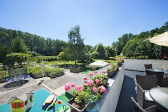 A spacious outdoor terrace featuring wicker furniture and a row of vibrant pink flowers in pots. In the foreground, there is a green table with toys and a small toy house. The scene overlooks a lush garden area, with tall trees lining the background and a trampoline visible on the patio. The sky is clear and blue, suggesting a sunny day.