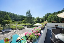 A spacious outdoor terrace featuring wicker furniture and a row of vibrant pink flowers in pots. In the foreground, there is a green table with toys and a small toy house. The scene overlooks a lush garden area, with tall trees lining the background and a trampoline visible on the patio. The sky is clear and blue, suggesting a sunny day.