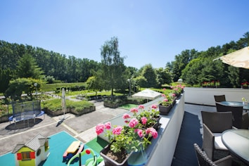 A spacious outdoor terrace featuring wicker furniture and a row of vibrant pink flowers in pots. In the foreground, there is a green table with toys and a small toy house. The scene overlooks a lush garden area, with tall trees lining the background and a trampoline visible on the patio. The sky is clear and blue, suggesting a sunny day.