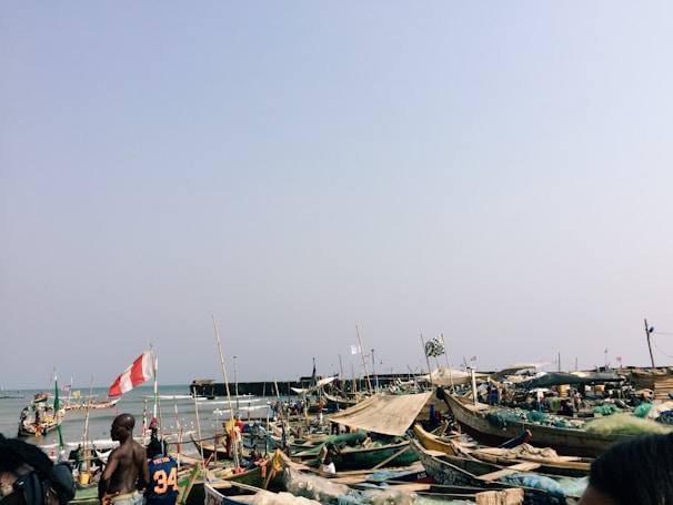 A coastal scene with numerous wooden boats lined up along the shore, many of which are adorned with flags. People are gathered around the boats, and some are looking out to the sea. The sea and sky take up a significant portion of the background, suggesting a sunny day.