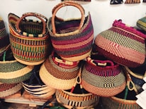 Colorful woven baskets are stacked together, each with unique patterns and vibrant hues. The baskets feature various designs, showcasing stripes and checkered patterns, with sturdy handles crafted from similar materials.