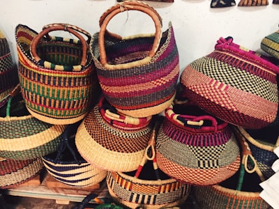 Colorful woven baskets are stacked together, each with unique patterns and vibrant hues. The baskets feature various designs, showcasing stripes and checkered patterns, with sturdy handles crafted from similar materials.