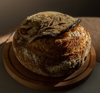 A rustic, round loaf of artisan bread rests on a wooden cutting board. The bread features a well-baked, crusty exterior with scoring patterns on top, and its warm brown tones are highlighted by soft, natural lighting, creating dramatic shadows.