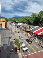 A bustling street scene in a small town with shops lining both sides of the road. Cars drive down the street and pedestrians walk on the sidewalks. Green trees and a cloudy sky serve as the backdrop. There are colorful storefronts and various people engaged in different activities.