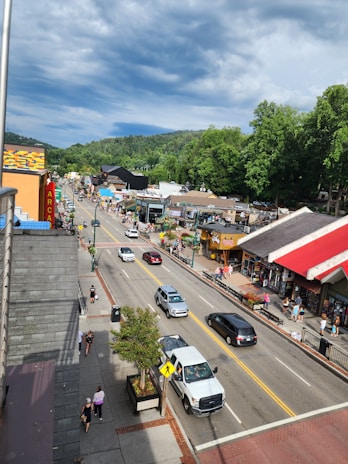 A bustling street scene in a small town with shops lining both sides of the road. Cars drive down the street and pedestrians walk on the sidewalks. Green trees and a cloudy sky serve as the backdrop. There are colorful storefronts and various people engaged in different activities.
