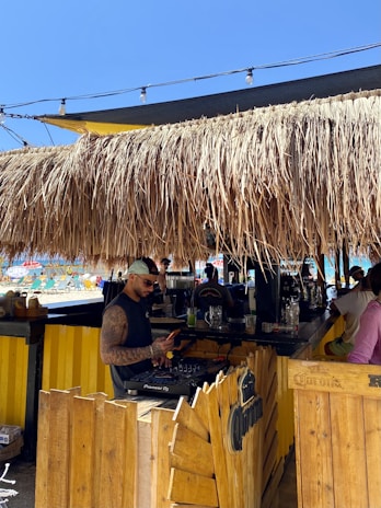A beachside bar with a thatched roof and a man standing at a DJ booth, browsing on a phone. The area is decorated in a tiki style with wooden elements and string lights overhead. In the background, there is a glimpse of the beach with people lounging under umbrellas.