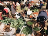 A bustling outdoor market scene with several women engaging in sorting and arranging a variety of fresh produce in woven baskets. The market is filled with vibrant vegetables like peppers, leafy greens, yams, and bananas. The women are dressed in brightly patterned clothing, reflecting a lively and colorful setting. The ground is covered with sacks and baskets, contributing to the busy marketplace atmosphere.