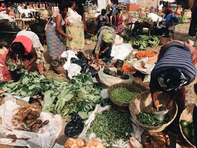 A bustling outdoor market scene with several women engaging in sorting and arranging a variety of fresh produce in woven baskets. The market is filled with vibrant vegetables like peppers, leafy greens, yams, and bananas. The women are dressed in brightly patterned clothing, reflecting a lively and colorful setting. The ground is covered with sacks and baskets, contributing to the busy marketplace atmosphere.