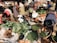 A bustling outdoor market scene with several women engaging in sorting and arranging a variety of fresh produce in woven baskets. The market is filled with vibrant vegetables like peppers, leafy greens, yams, and bananas. The women are dressed in brightly patterned clothing, reflecting a lively and colorful setting. The ground is covered with sacks and baskets, contributing to the busy marketplace atmosphere.