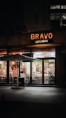 A warmly lit restaurant facade with the sign 'BRAVO EAT & DRINK' illuminated in orange-yellow light. The storefront features large glass windows showing the interior, with tables and chairs arranged neatly. An outdoor seating area is defined by a prominent large white umbrella. Ambient lighting creates a cozy and inviting atmosphere, with a few subtle reflections on the windows.