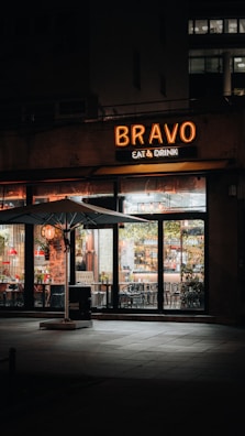 A warmly lit restaurant facade with the sign 'BRAVO EAT & DRINK' illuminated in orange-yellow light. The storefront features large glass windows showing the interior, with tables and chairs arranged neatly. An outdoor seating area is defined by a prominent large white umbrella. Ambient lighting creates a cozy and inviting atmosphere, with a few subtle reflections on the windows.
