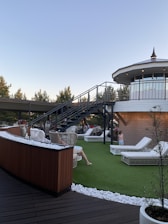 A rooftop terrace with wicker chairs and white lounge beds on artificial grass is depicted. A modern staircase leads up to an enclosed, circular structure with large windows. People are relaxing on the terrace under a clear sky, surrounded by trees and decorative white stones.