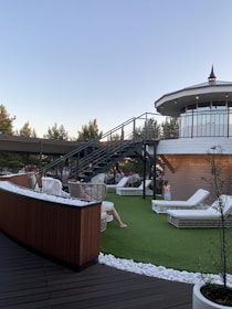A rooftop terrace with wicker chairs and white lounge beds on artificial grass is depicted. A modern staircase leads up to an enclosed, circular structure with large windows. People are relaxing on the terrace under a clear sky, surrounded by trees and decorative white stones.