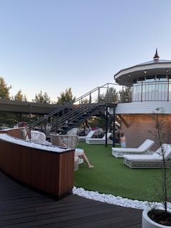 A rooftop terrace with wicker chairs and white lounge beds on artificial grass is depicted. A modern staircase leads up to an enclosed, circular structure with large windows. People are relaxing on the terrace under a clear sky, surrounded by trees and decorative white stones.