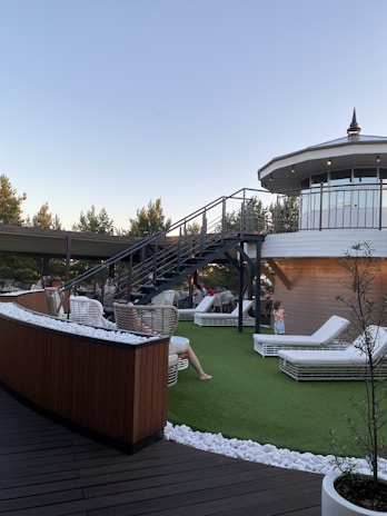 A rooftop terrace with wicker chairs and white lounge beds on artificial grass is depicted. A modern staircase leads up to an enclosed, circular structure with large windows. People are relaxing on the terrace under a clear sky, surrounded by trees and decorative white stones.