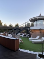 A rooftop terrace with wicker chairs and white lounge beds on artificial grass is depicted. A modern staircase leads up to an enclosed, circular structure with large windows. People are relaxing on the terrace under a clear sky, surrounded by trees and decorative white stones.
