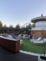 A rooftop terrace with wicker chairs and white lounge beds on artificial grass is depicted. A modern staircase leads up to an enclosed, circular structure with large windows. People are relaxing on the terrace under a clear sky, surrounded by trees and decorative white stones.