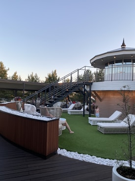 A rooftop terrace with wicker chairs and white lounge beds on artificial grass is depicted. A modern staircase leads up to an enclosed, circular structure with large windows. People are relaxing on the terrace under a clear sky, surrounded by trees and decorative white stones.