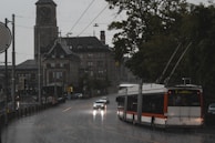 A city street scene during rainfall with a public trolleybus and a car driving along a wet road. In the background, there is a large historical building with a clock tower, and to the right, there are trees lining the street.