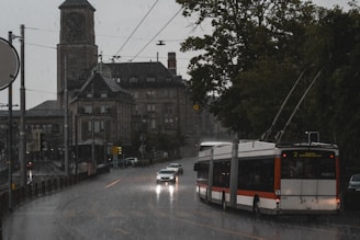 A city street scene during rainfall with a public trolleybus and a car driving along a wet road. In the background, there is a large historical building with a clock tower, and to the right, there are trees lining the street.