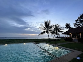 A luxurious coastal-style pool area with natural wood textures, bright green foliage, and subtle sky-blue lighting at dusk.