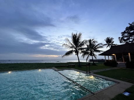A luxurious coastal-style pool area with natural wood textures, bright green foliage, and subtle sky-blue lighting at dusk.