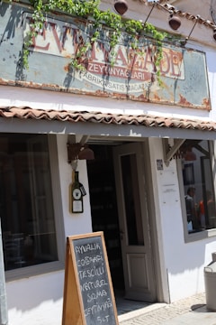 An exterior view of a rustic shop with a weathered sign, surrounded by green vines, above the entrance. Below the sign, there is a large window and a door, both framed in neutral colors. In front of the shop, a wooden chalkboard sign displays text in a foreign language. The building has a tiled roof edge, and sunlight casts shadows on the facade.