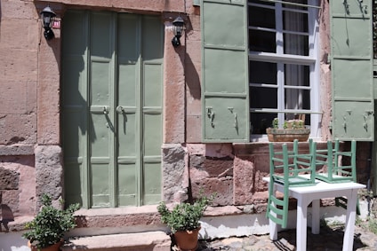 The image shows a charming exterior of a rustic building with stone walls. There is a green door with matching green window shutters. In front of the window, there is a small white table with two green chairs placed on top. Potted plants are positioned beside the door. Two wall-mounted lamps flank the door.