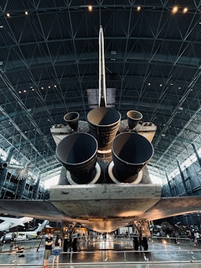 A large spacecraft is displayed inside a spacious museum hangar, showcasing its massive engines from a rear view. The interior architecture features a high ceiling with geometric patterns, and visitors are seen observing the exhibit.