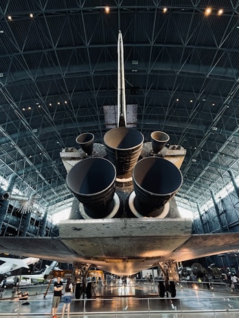 A large spacecraft is displayed inside a spacious museum hangar, showcasing its massive engines from a rear view. The interior architecture features a high ceiling with geometric patterns, and visitors are seen observing the exhibit.