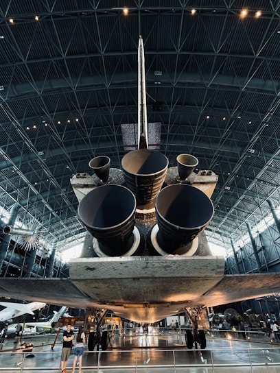 A large spacecraft is displayed inside a spacious museum hangar, showcasing its massive engines from a rear view. The interior architecture features a high ceiling with geometric patterns, and visitors are seen observing the exhibit.
