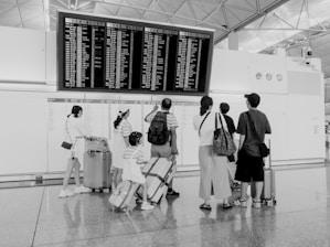 A group of people stand in front of a large flight information board at an airport. They are looking at the departure and arrival details. The scene is monochrome, and several individuals have luggage with them. The architecture of the space includes high ceilings and industrial elements.